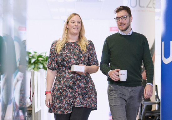 Two people walking through a modern office corridor while carrying mugs, with desks, office furniture, glass partitions, and workplace branding visible in the background.