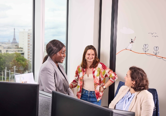 Three people in conversation within a modern office workspace, standing and seated beside desks and computer monitors. Large windows overlook a cityscape in the background, while wall illustrations and collaborative workspace features are visible behind them