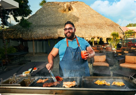 Person grilling food outdoors while holding a colorful drink.